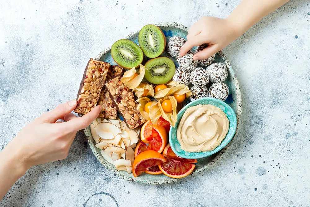 hands-grabbing-snacks-bars-hummus-fruit-nuts-on-bowl-plate-blue-white-background